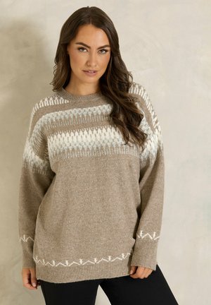 Femme aux longs cheveux bruns portant un pull beige avec des motifs géométriques blancs et marron clair, debout devant un fond neutre.