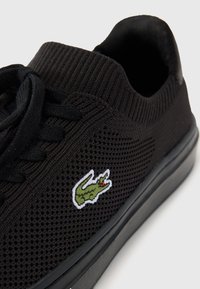 Sneaker nera in rete con colletto rigato, lacci neri piatti e logo di coccodrillo verde ricamato sul lato. Suola in gomma.