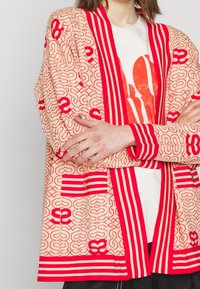 Personne portant un cardigan beige et rouge à motifs avec des rayures audacieuses par-dessus une chemise blanche avec un grand texte graphique orange. Bras croisés.