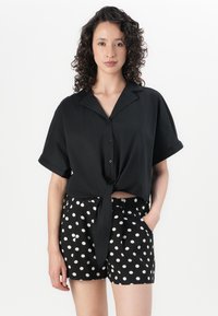 Crna majica s kratkim rukavima s detaljem vezanja sprijeda, u kombinaciji s crnim hlačama visokog struka koje imaju bijele točkice i bočne džepove.