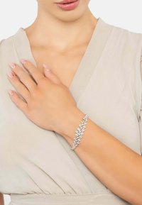 Woman wearing a teardrop-shaped crystal bracelet on her wrist, dressed in a light beige V-neck garment, with manicured nails.