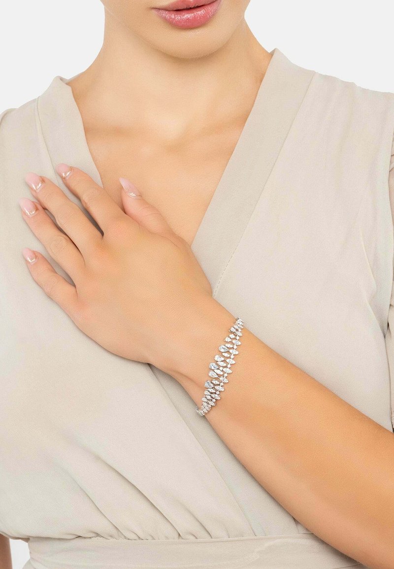 Woman wearing a teardrop-shaped crystal bracelet on her wrist, dressed in a light beige V-neck garment, with manicured nails.