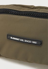 Zielona oliwkowa torba z tkaniny ma czarny zamek błyskawiczny oraz teksturowaną czarną metkę z białym tekstem "ELEMENT CO. SINCE 1992." Prosty i funkcjonalny design.