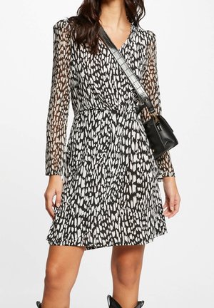 Kobieta ubrana w czarno-białą sukienkę kopertową z przezroczystymi długimi rękawami, nosząca czarną torebkę na ramię z szerokim paskiem.