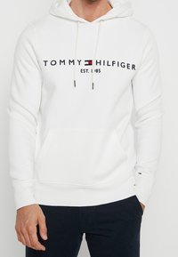 Bílá mikina s kapucí vyrobená z měkké látky. Má přední kapsu, šňůrky na stažení a černé potiskové logo s červeným a modrým akcentem.