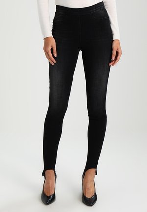 Woman wearing black skinny jeans and black pointed heels, standing with hands relaxed at sides against plain background.