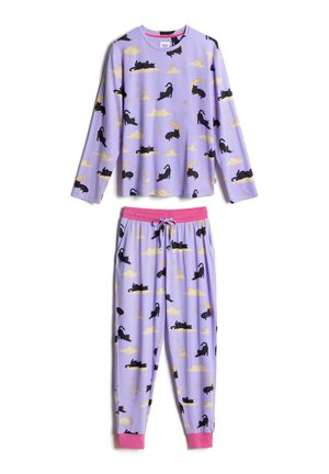 Mujer vestida con pijama lavanda a juego con patrones de gato negro, luna y estrellas, con las manos en los bolsillos, de pie frente a un fondo liso.
