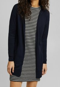 Lange marineblauwe cardigan met open voorkant, ribbelstructuur en zijkanten met zakken, gedragen over een zwart-wit gestreepte jurk.