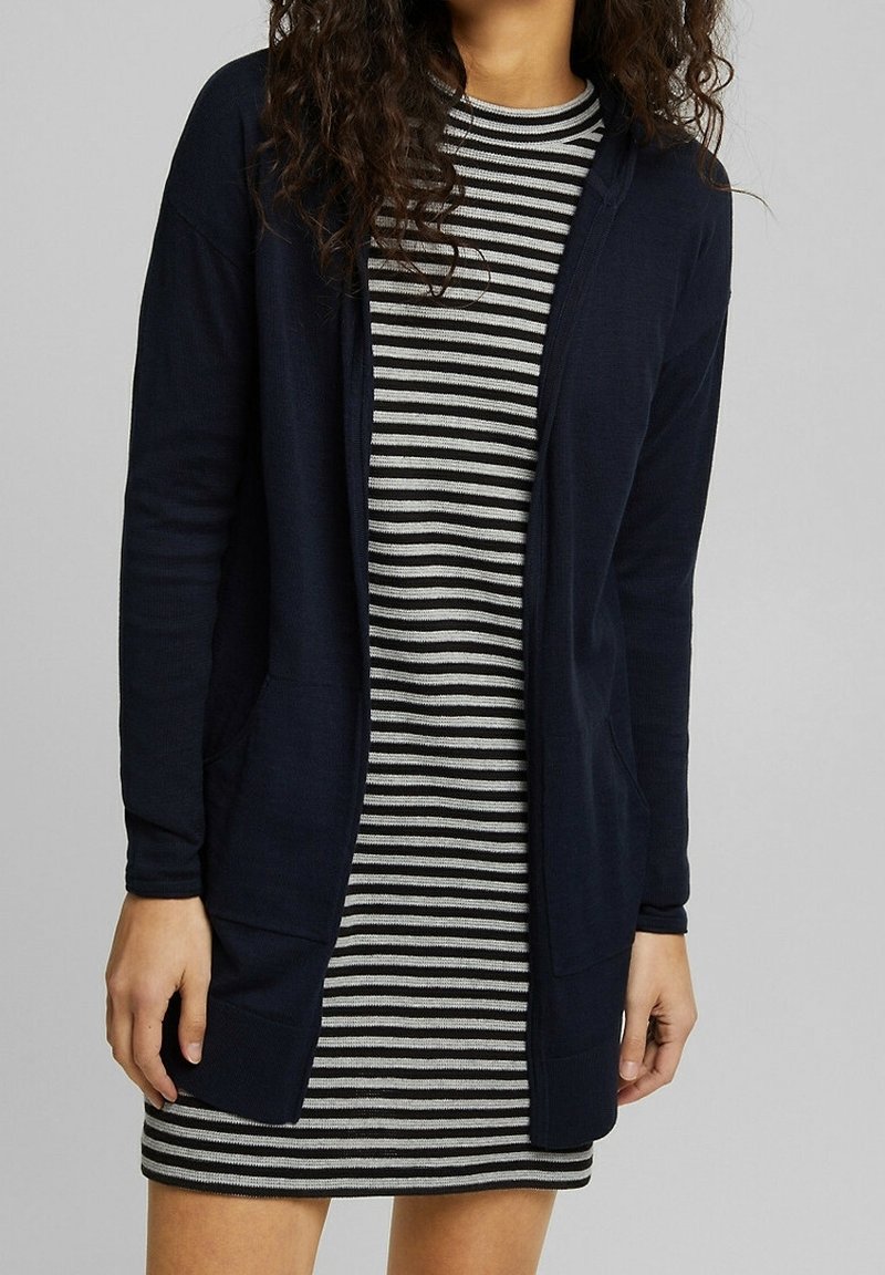 Lange marineblauwe cardigan met open voorkant, ribbelstructuur en zijkanten met zakken, gedragen over een zwart-wit gestreepte jurk.