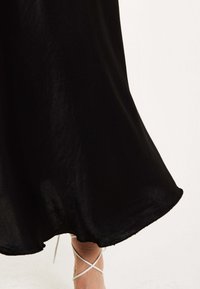 Black, flowing dress with a smooth texture, featuring a hemline that curves gently. White strappy sandals visible at the bottom.