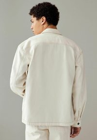 Chaqueta de mezclilla beige con un corte relajado, detalles de costura visibles, puños abotonados y un diseño liso en la parte trasera.