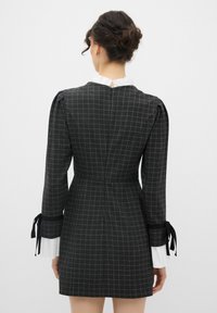 Sort ternet kjole med et tætsiddende design, puffede ærmer med bindebånd ved manchetten, og en høj hvid krave. Lynlås lukning bagpå er synlig.