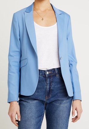 Vrouw draagt een lichtblauwe blazer over een wit shirt, gecombineerd met hooggetailleerde blauwe jeans en laagjes gouden kettingen.