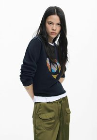 Granatowa bluza z kolorową haftem i ściągaczami na rękawach, noszona na białej koszuli, zestawiona z oliwkowozielonymi satynowymi spodniami z dużymi kieszeniami.