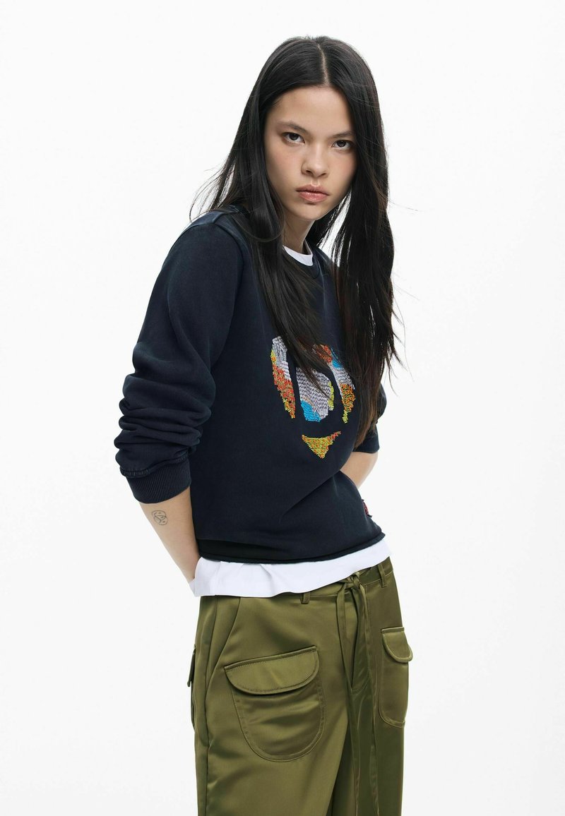 Granatowa bluza z kolorową haftem i ściągaczami na rękawach, noszona na białej koszuli, zestawiona z oliwkowozielonymi satynowymi spodniami z dużymi kieszeniami.