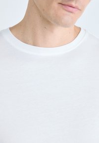 Gros plan sur le cou et le bas du visage d'une personne portant un t-shirt blanc uni à col rond, sur fond clair.