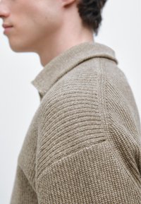 Pull beige en tricot avec un motif texturé, doté d'un col côtelé et d'une silhouette ajustée, présenté de profil.