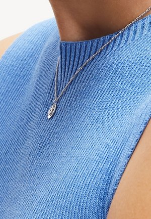 Gros plan sur une personne portant un collier pendentif en argent et un haut tricoté sans manches bleu clair avec un col côtelé.