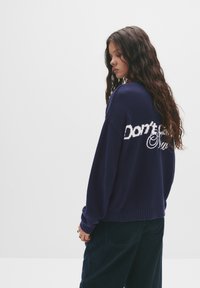 Tengerészkék kötött pulóver, a hátsó részén fehér "Don't" felirattal, laza szabással és bordázott szegéllyel. Sötét corduroy nadrággal párosítva.