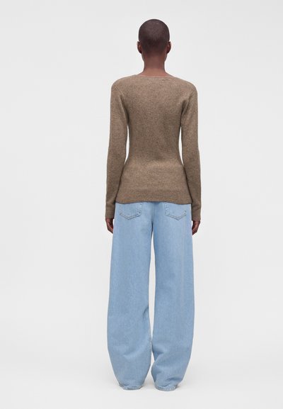 Personne debout, de dos, portant un haut marron moulant à manches longues et un jean bleu clair ample sur un fond uni.