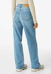 Person klädd i ljusblå hög midja raka jeans och en beige långärmad topp, stående med ena handen avslappnad vid sidan.