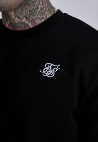 Camiseta de algodón negra con un pequeño logo bordado en blanco. Cuello redondo, textura suave y diseño ajustado.