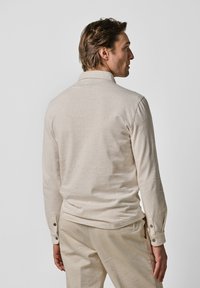 Beige långärmad polo med mjukt tyg. Har en krage, knappdetaljer vid ärmsluten och en avslappnad passform.