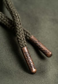 Geflochtene olivgrüne Schnürsenkel mit bronzefarbenen Schuhspitzen, die ein graviertes Logo zeigen. Glatte Textur vor einem Stoffhintergrund.