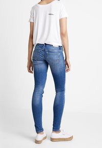 Skinny blue denim jeans with a mid-rise waist, worn with a white short-sleeve shirt and white sneakers. Features a faded wash and two back pockets.