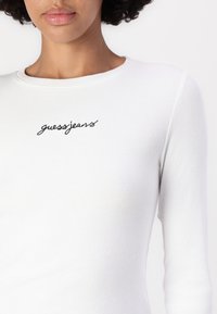 Biely rebrovaný top s dlhými rukávmi, na ktorom je na hrudi vyšívané čierne logo "guessjeans". Hladká textúra a priliehajúci strih.