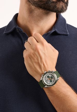 Montre chronographe en argent avec bracelet en silicone vert, cadran blanc, accents noirs et sous-cadrans. Portée sur une main sur un fond de polo bleu marine.