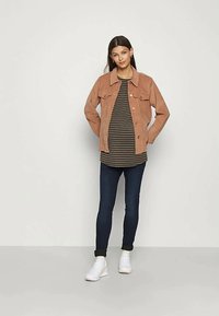 Mulher está encostada numa parede lisa, vestindo um casaco de corduroy castanho, uma camisa listrada preta e castanha, jeans escuros e ténis brancos, com as mãos atrás das costas.