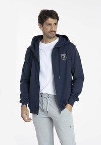 Sweatshirt zippé bleu marine avec capuche, arborant un patch logo sur le devant, associé à un pantalon de survêtement gris clair à cordon. Texture de tissu lisse.