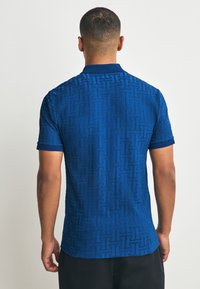 Polo met korte mouwen in diepblauw stof met subtiel tonale geometrische patroon. Voorzien van een klassieke kraag en contrasterende textuur op de mouwboorden.