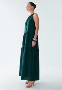 Žalios spalvos ilga be rankovių suknelė laisvo silueto, sluoksniuota konstrukcija ir minkšta medžiaga, derinama su kūno spalvos basutėmis su dirželiais ir minimalistiniais aksesuarais.