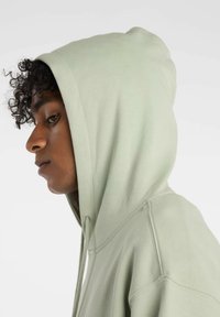 Felpa verde chiaro con una texture morbida e cappuccio con lacci, caratterizzata da una vestibilità comoda e dettagli di cucitura visibili.