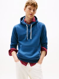 Sudadera azul con un bolsillo frontal y cordones, superpuesta sobre una camiseta de rayas rojas. Pantalones beige completan el atuendo.