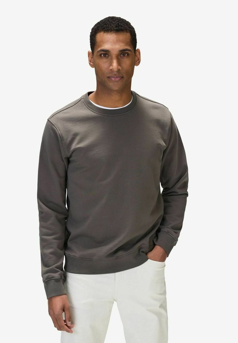 Mörkgrå sweatshirt med rund hals, ribbade muddar och en slät textur, bärs över vita byxor av en person som står med händerna i fickorna.