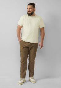 Kremowa koszulka z krótkim rękawem w połączeniu z brązowymi spodniami cargo i beżowymi sneakersami. Prosty krój z luźnym fasonem i widocznymi detalami przeszycia.