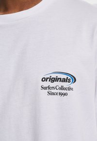 Hvit bomulls t-skjorte med rund hals, med en svart og blå logo trykt på brystet som leser "originals Surfers Collective Since 1990."