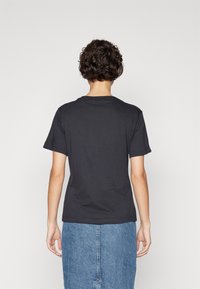 Person med kort krøllet hår iført en ensfarvet sort t-shirt og en blå denimnederdel, stående med ansigtet vendt mod en hvid baggrund.