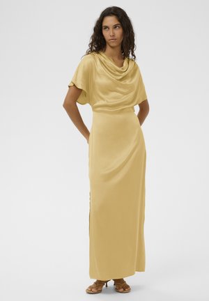 Kobieta o kręconych włosach, ubrana w musztardowożółtą satynową suknię maxi z dekoltem typu drapowanie i sandałami, stojąca z rękami za plecami.