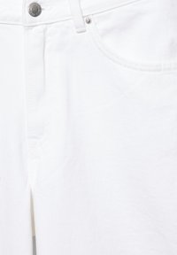 Gros plan sur un jean en denim blanc montrant la poche avant, la boucle de ceinture et un bouton en métal avec gravure de la marque.