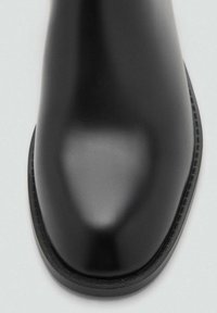 Botina din piele neagră cu vârful rotunjit, finisaj neted și un detaliu subtil de cusătură de-a lungul marginii tălpii. Design cu toc plat.