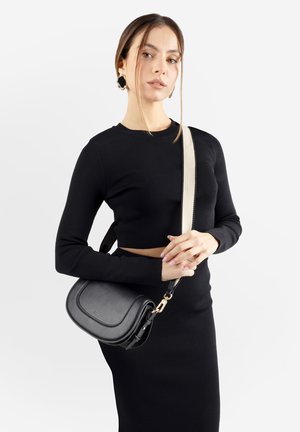 Donna che indossa una maglia aderente a maniche lunghe nera e una gonna, con una borsa a tracolla nera con tracolla color crema, in piedi su uno sfondo bianco.