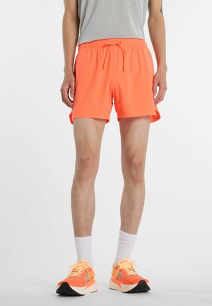 Person iført lyse orange atletikshorts, grå t-shirt, hvide crew-sokker og orange løbesko, står mod en ensfarvet baggrund.