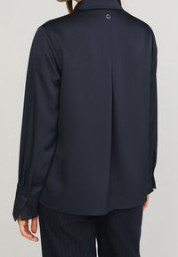 Kobieta w granatowej bluzce z długim rękawem, z plisą z tyłu i zapięciem na guziki przy mankietach, zestawiona z ciemnymi spodniami w prążki.