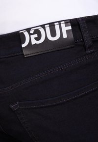 Jeans in denim scuro con un'etichetta in pelle che mostra "HUGO" in grande font bianco, evidenziando il cinturino e i dettagli di cucitura eleganti.