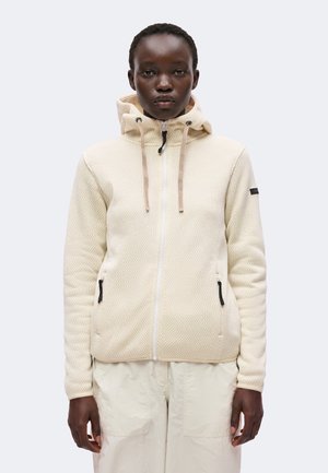 Person iført en lys beige tekstureret lynlåstrøje med hætte og beige snore, kombineret med off-white bukser mod en ensfarvet baggrund.