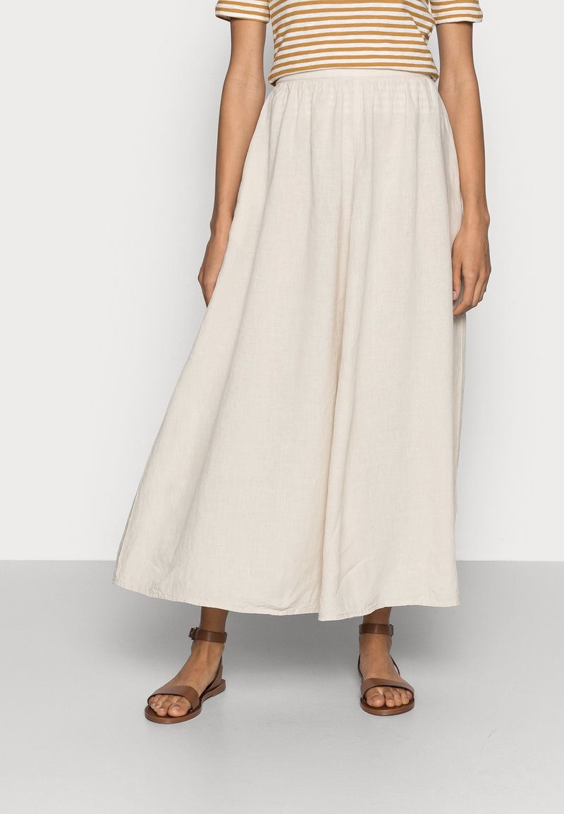 Lange, vloeiende beige rok gemaakt van lichte stof met een geplooide tailleband. Gepaard met bruine platte sandalen en een gestreepte top.